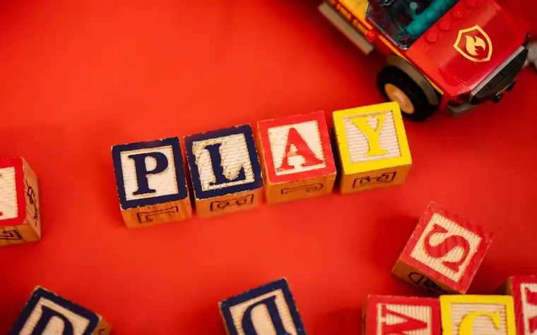 빨간 바닥 위에 알파벳 나무 블록으로 만든 'PLAY' 글자와 장난감 소방차
